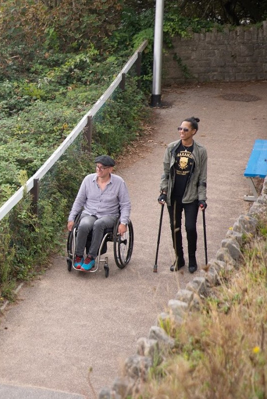 Two people are on a path near some trees. One is wearing glasses and using a wheelchair; the other is using a stick.